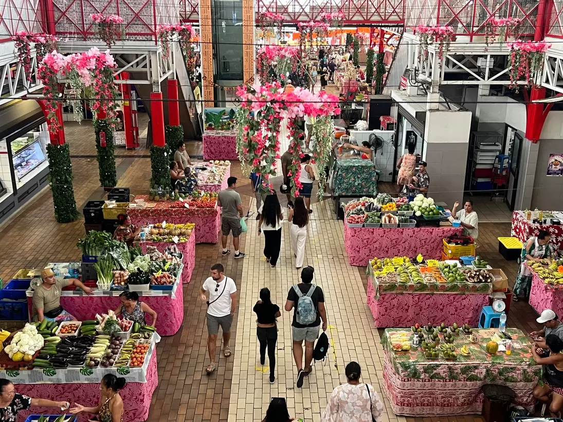 Market hall seen from above, fruit and vegetable tables draped in pink floral cloth, shoppers in the aisle.