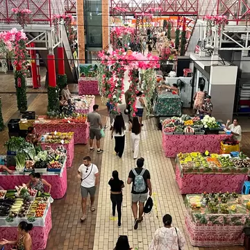 Market hall seen from above, fruit and vegetable tables draped in pink floral cloth, shoppers in the aisle.