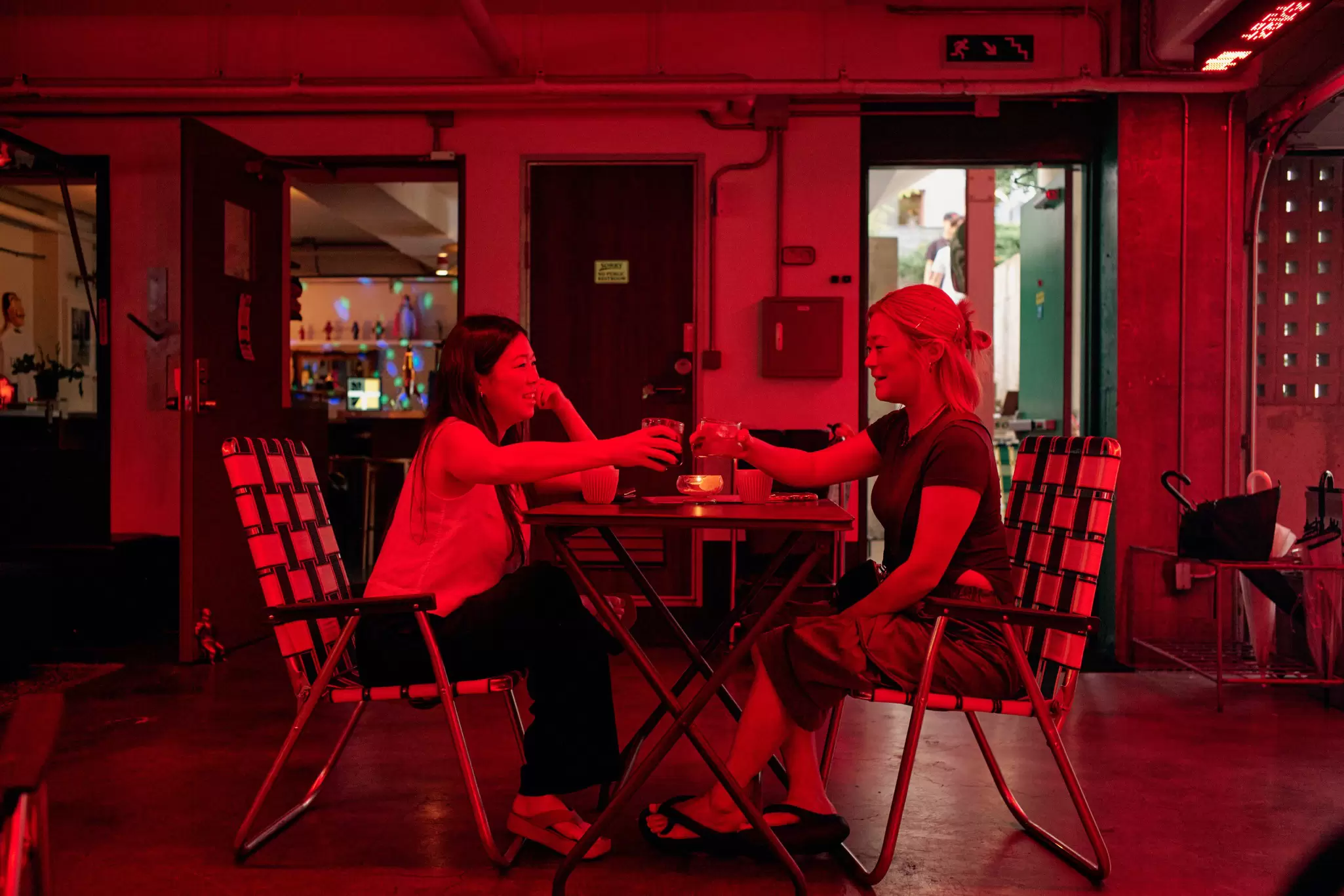 Two women toast glasses at an outdoor table bathed in red light.