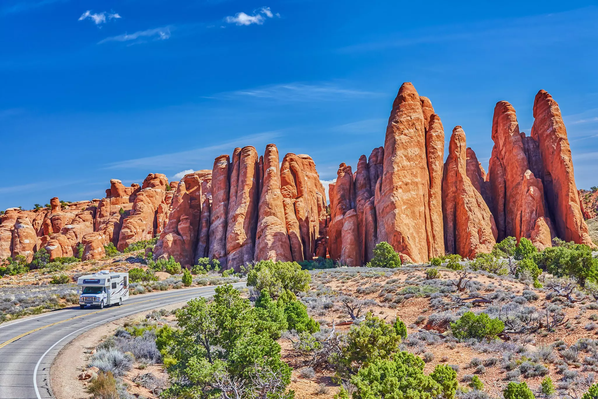 An RV drives on a two-lane blacktop road around a curve with reddish rock formations behind it; the sky is blue, and there are green bushes to the side of the road.