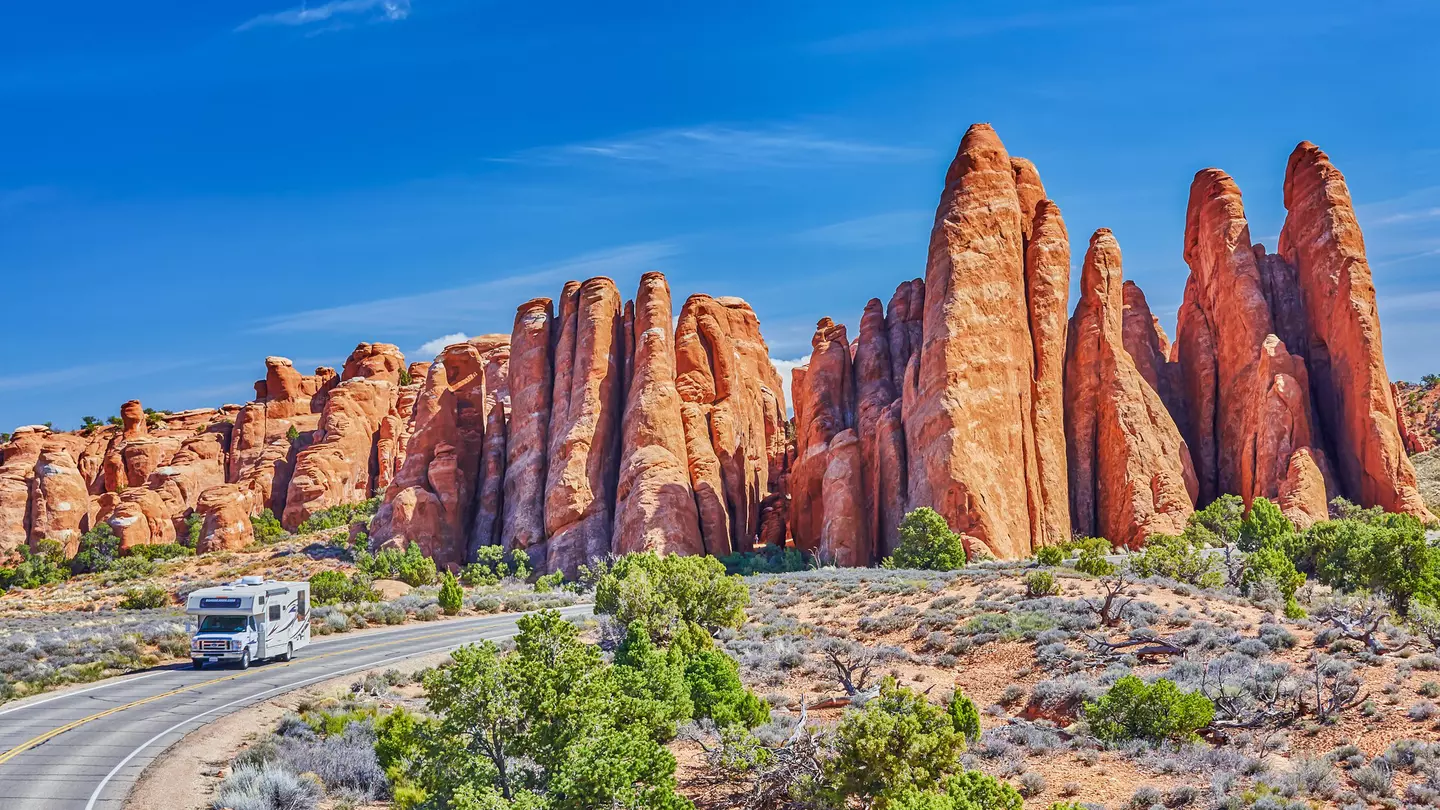An RV drives on a two-lane blacktop road around a curve with reddish rock formations behind it; the sky is blue, and there are green bushes to the side of the road.