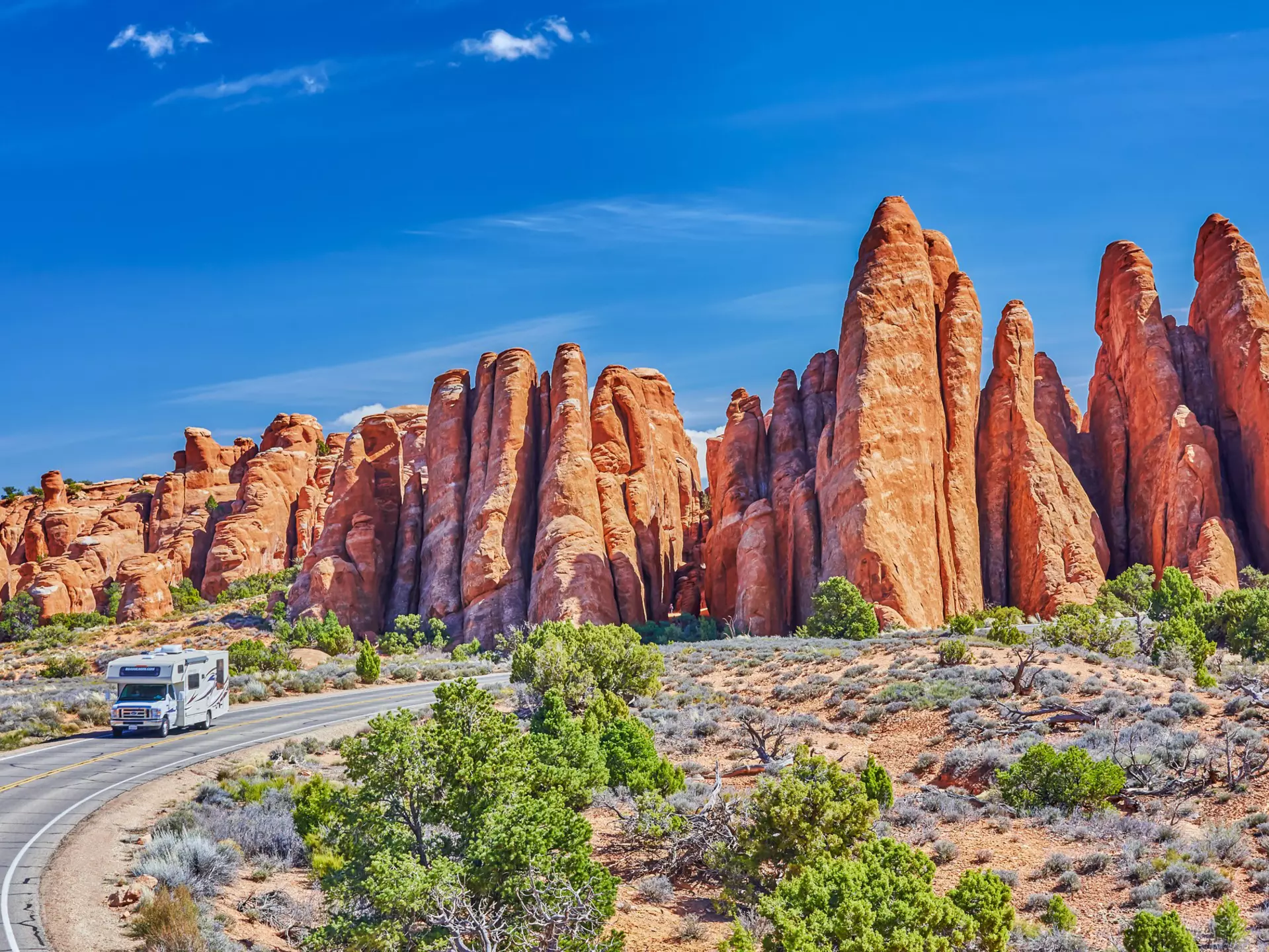 An RV drives on a two-lane blacktop road around a curve with reddish rock formations behind it; the sky is blue, and there are green bushes to the side of the road.