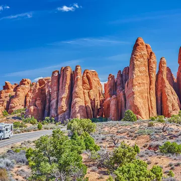 An RV drives on a two-lane blacktop road around a curve with reddish rock formations behind it; the sky is blue, and there are green bushes to the side of the road.
