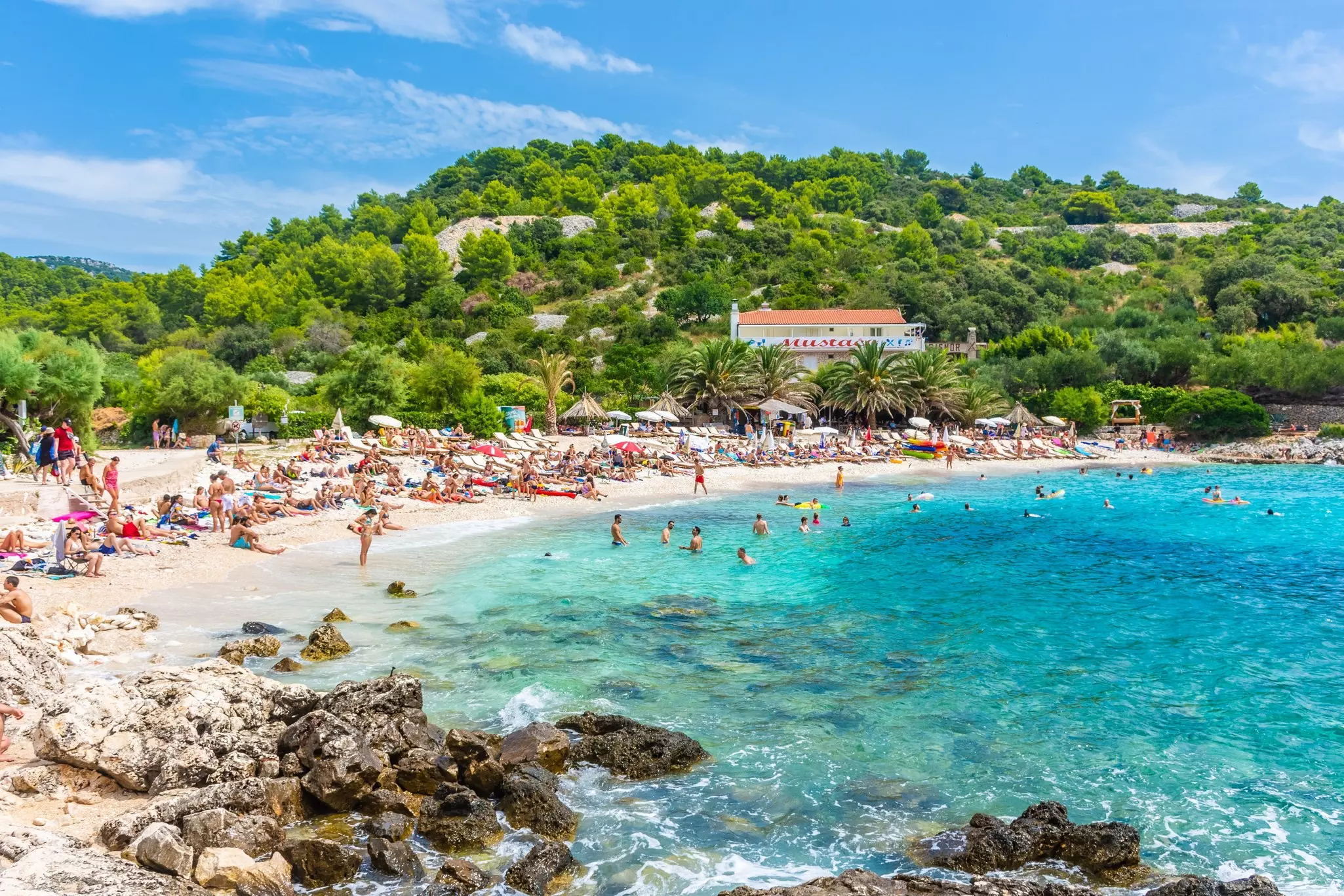 People swimming and sunbathing on a busy beach framed by trees and hills.