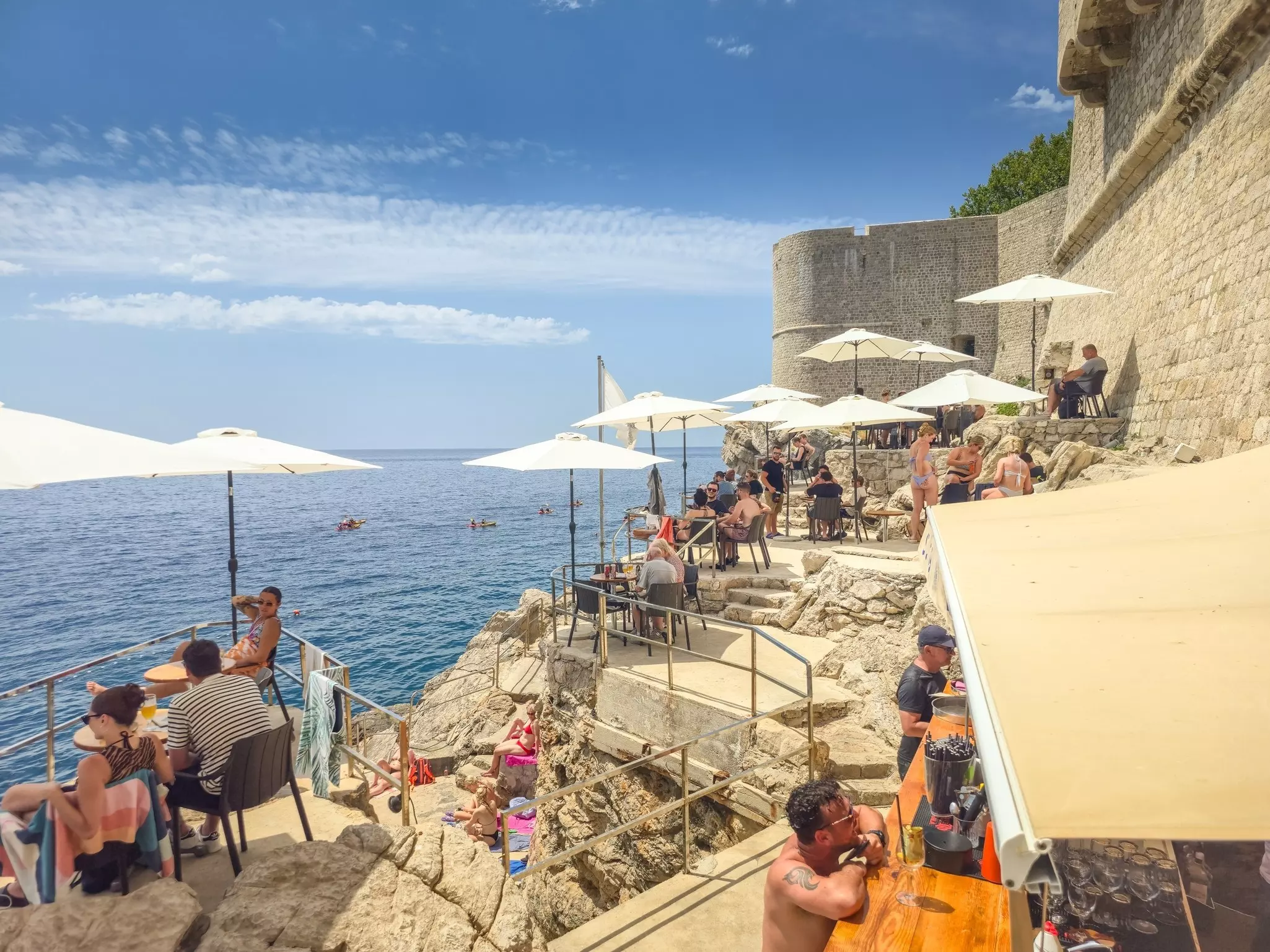 People sit at tables under sunshades at a bar on rocky cliffs at the edge of the sea.