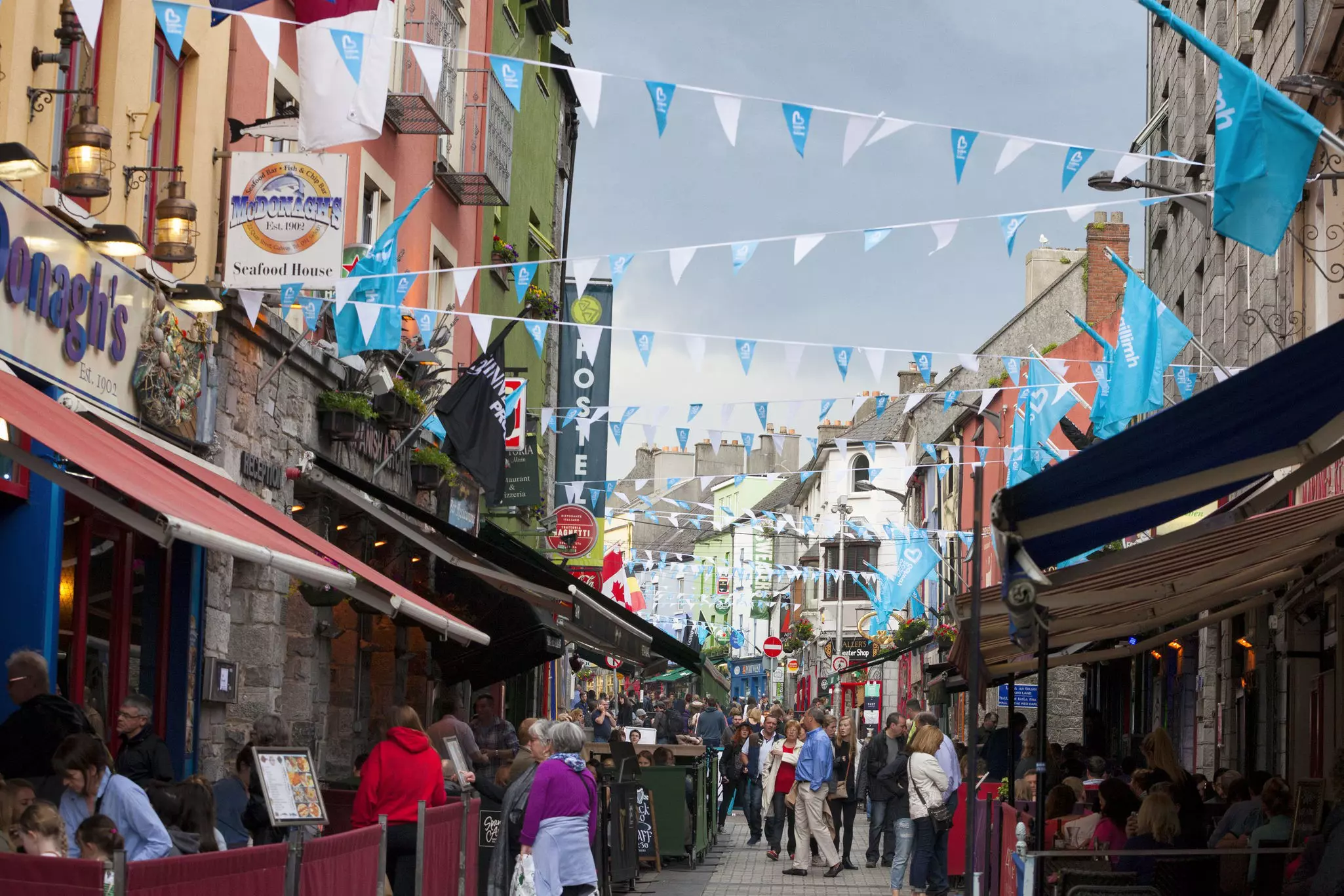 People walk down a crowded historic street lined with shops and bars. Pennants zig zag between the buildings above the street. Galway, Ireland.