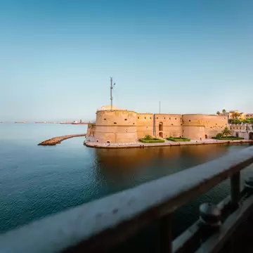 A round-towered stone castle on the edge of calm blue water in Taranto, Italy, with an iron railing in the foreground.