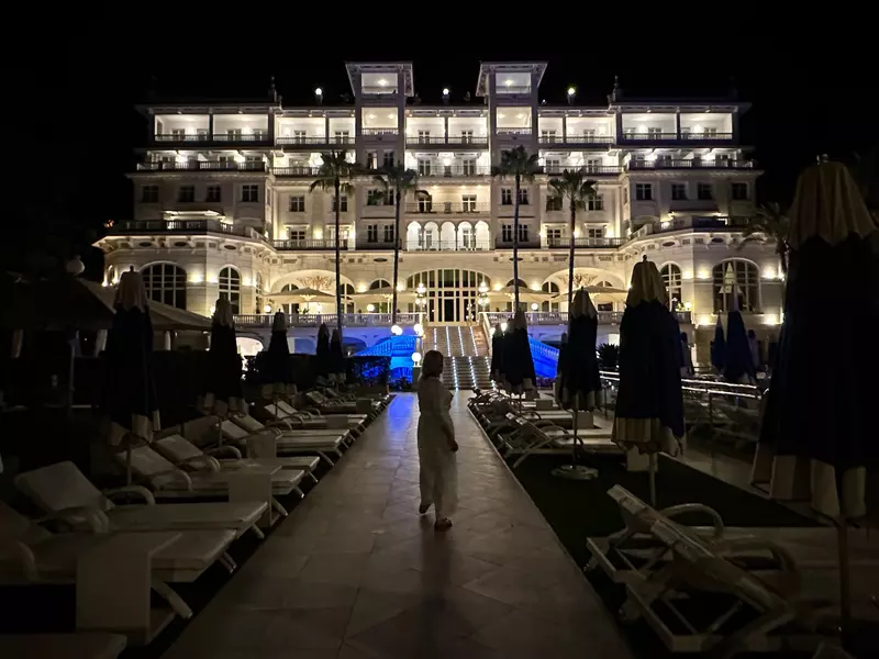 A person on a walkway between empty lounge chairs; a white building lit up at night is in the background.