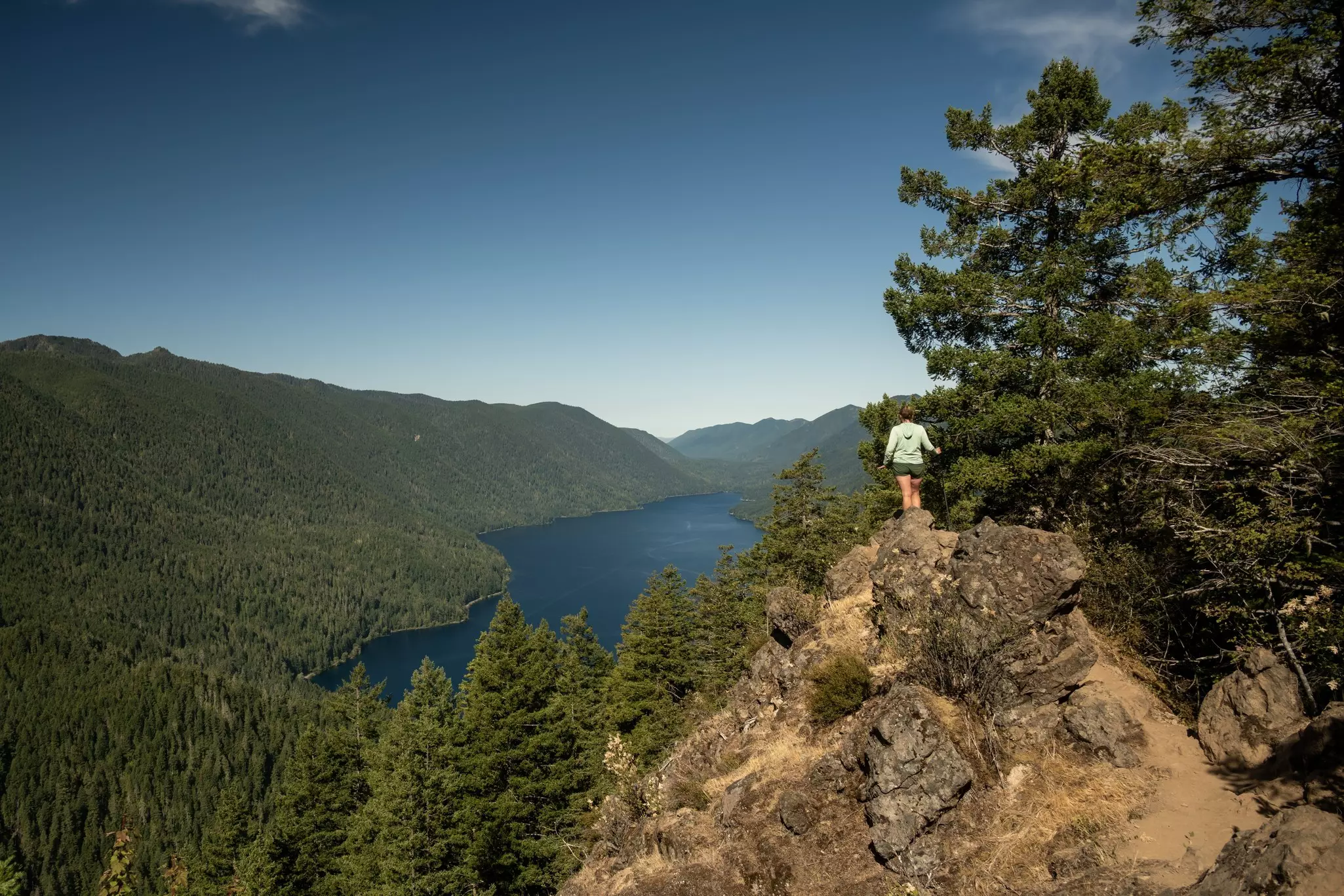 A hiker stands on top of rocks, looking down at a lake surrounded by green trees.