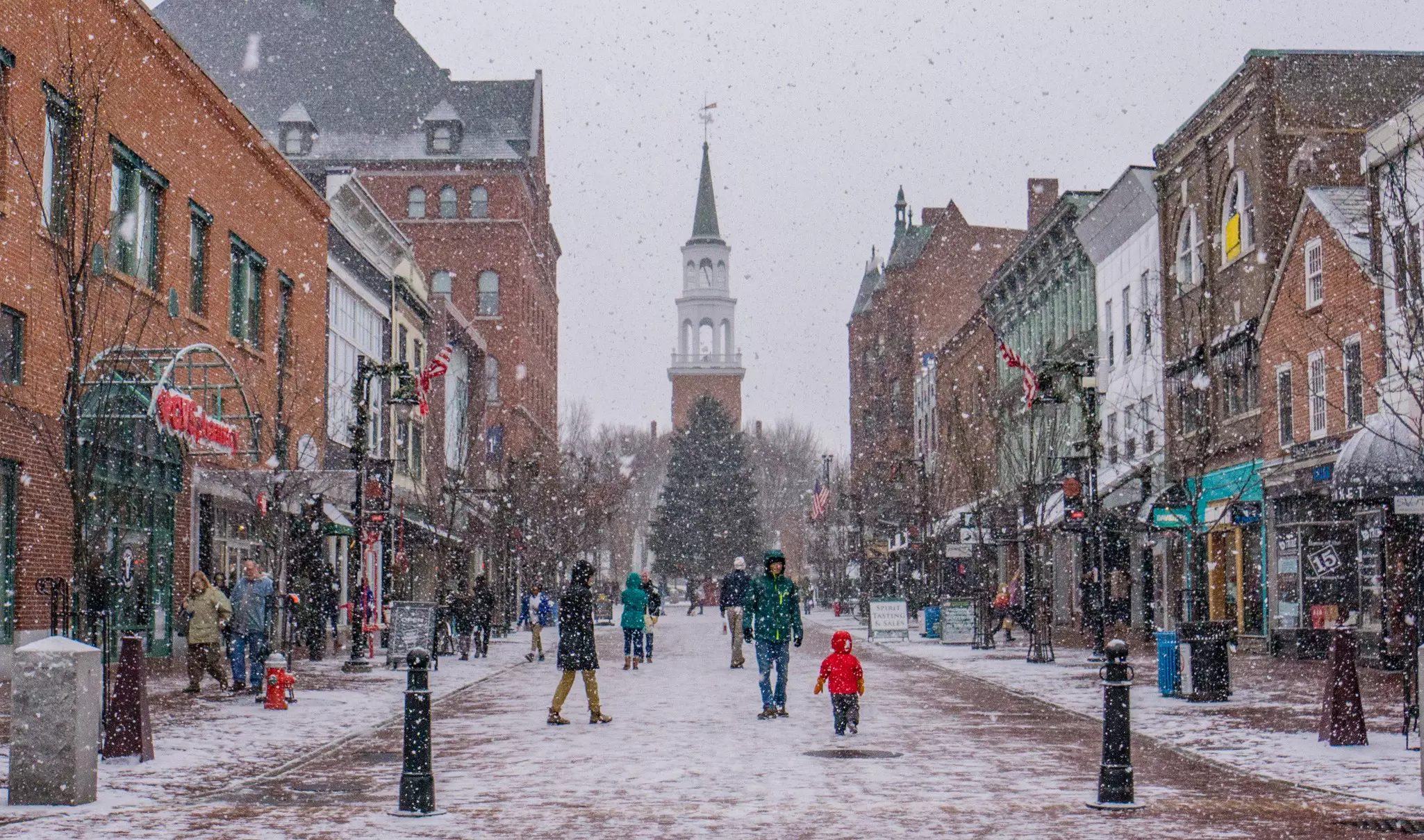 People walking through snow flurries on a picturesque New England pedestrian plaza where brick buildings are sandwiched together along the way