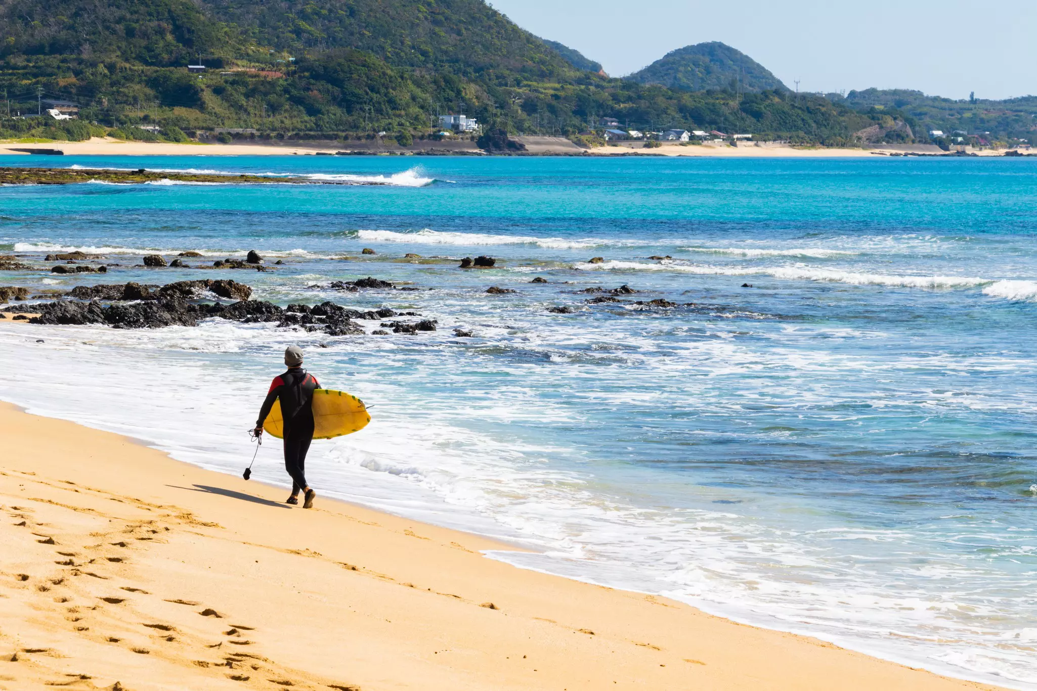 A person carried a surfboard along a sandy beach with waves crashing on the shore in the distance.