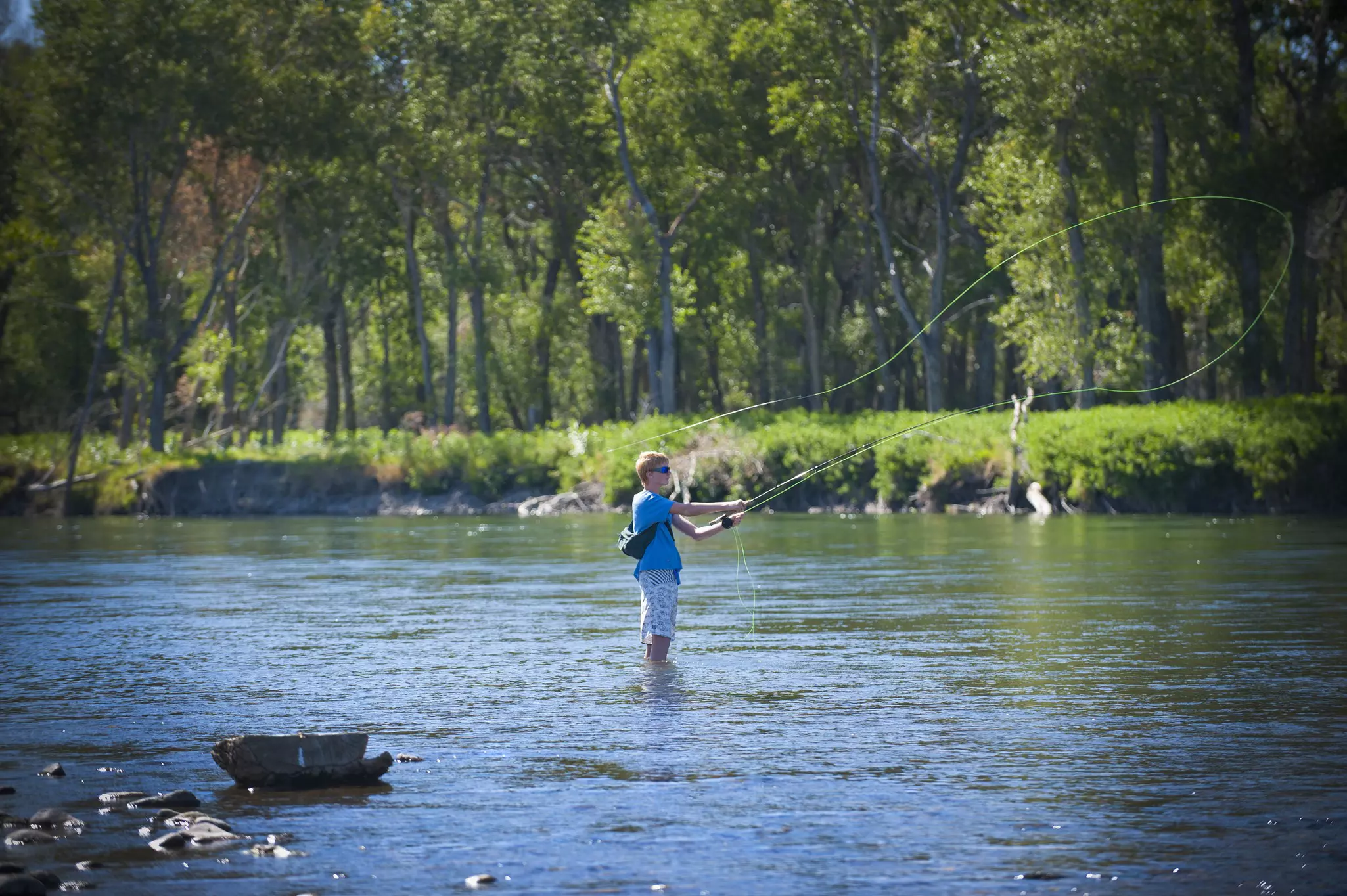 A person stands knee-deep in a river with a fishing rod on a sunny day.