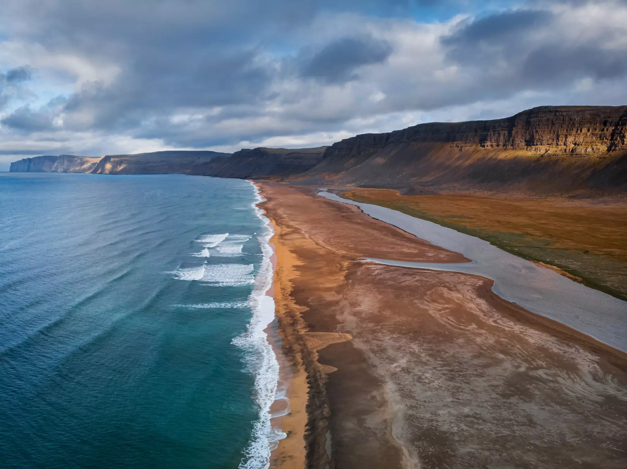 An aerial view of waves hitting a beach in the Westfjords, Iceland. The sand on the beach appears rust colored.