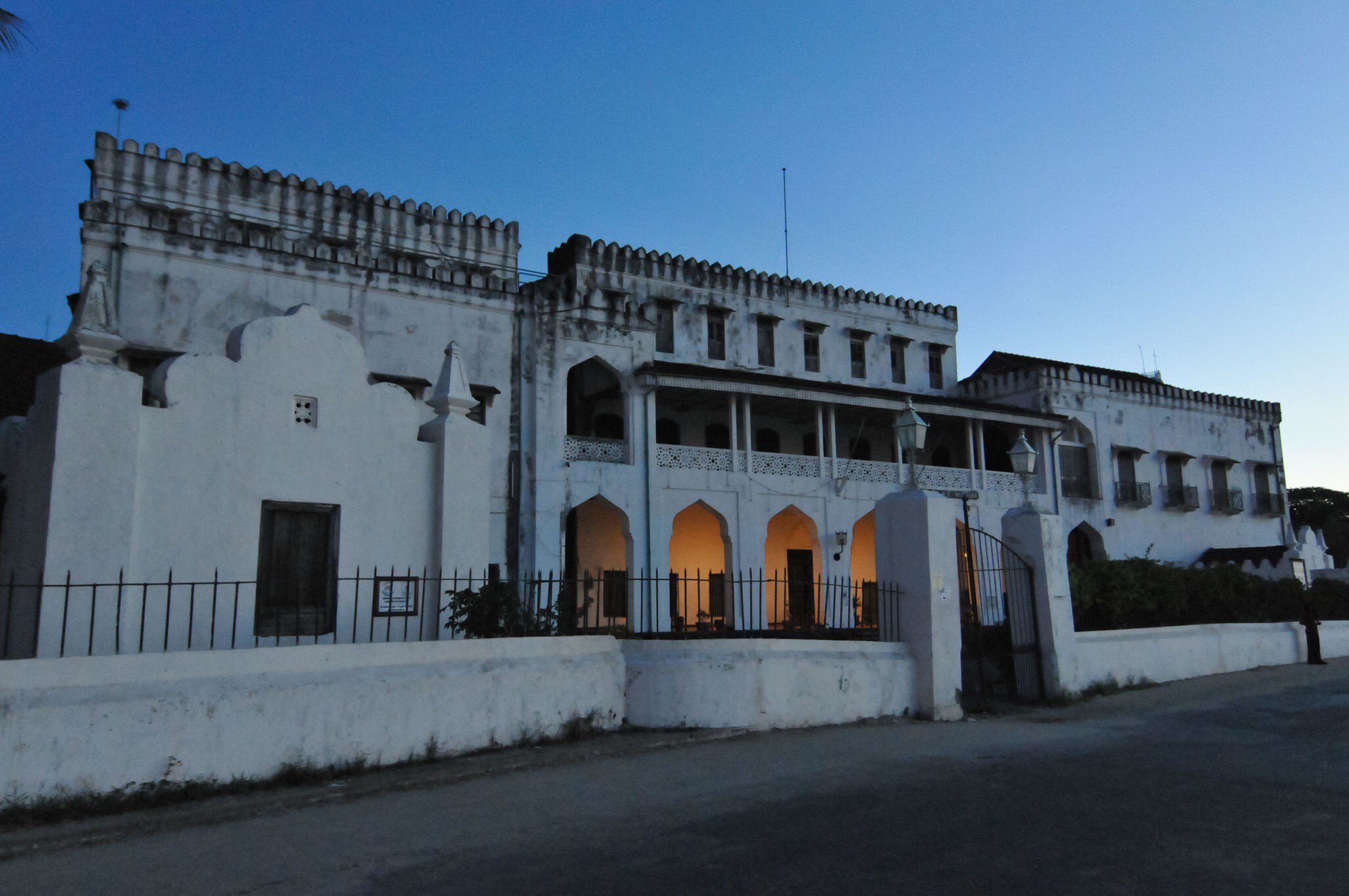 A whitewashed palace with crenellated parapets and a pillared verandah at dusk, warm light glowing through its ground-floor arches.