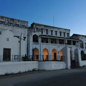 A whitewashed palace with crenellated parapets and a pillared verandah at dusk, warm light glowing through its ground-floor arches.