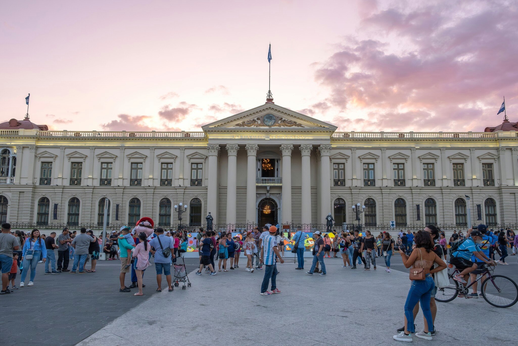 Many Salvadoran people and tourists walking at plaza in historical city center main square in front of National Palace. Beautiful sunset clouds in background.