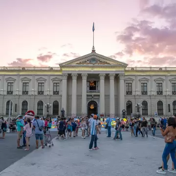 Many Salvadoran people and tourists walking at plaza in historical city center main square in front of National Palace. Beautiful sunset clouds in background.