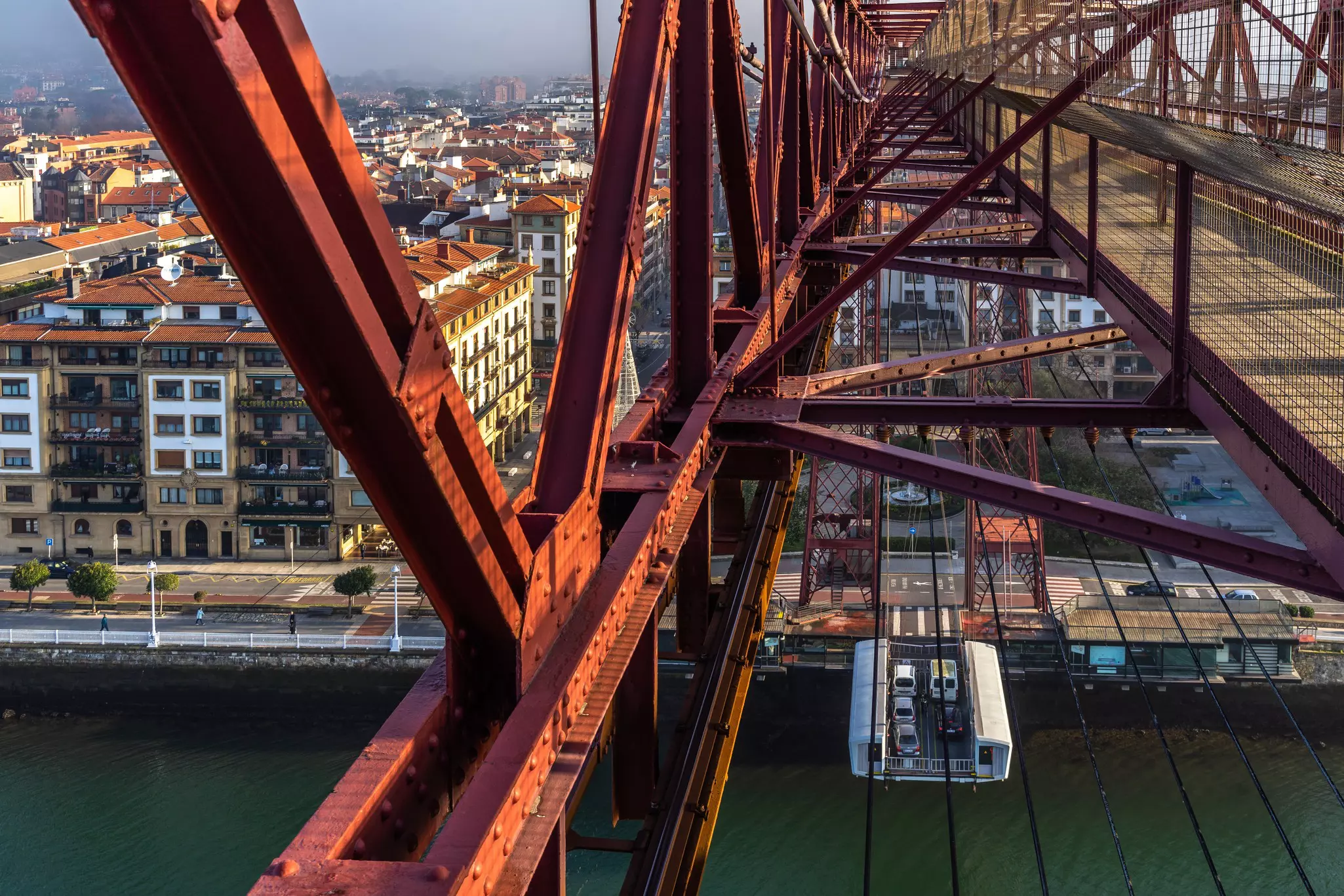 A view from a bridge in a city reveals its red trusses, and a walkway high above. A gondola carries cars below.