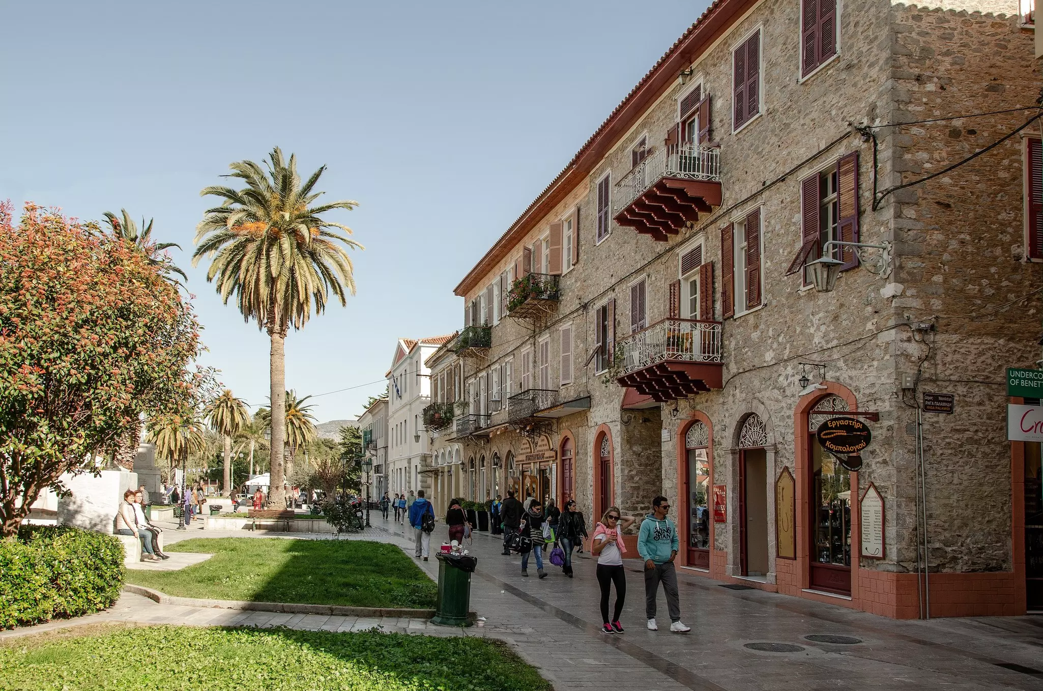 People walk past stone buildings next to a park in a city.