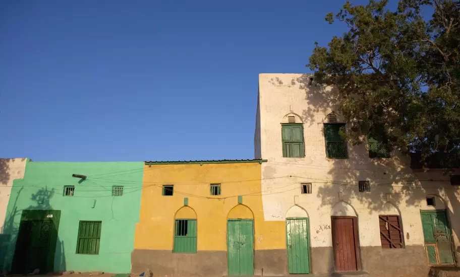 Row of mint-green, yellow and whitewashed houses with green shutters and arched doorways on a sandy street, a tree at right