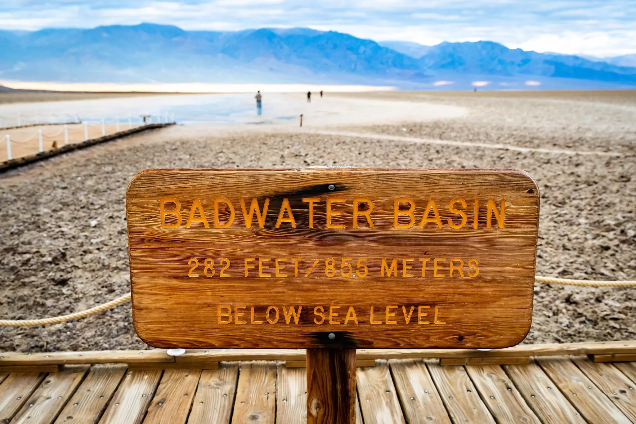 A wooden sign in front of a salt flat where people are walking says "Badwater Basin, 282 feet/855 metres below sea level"