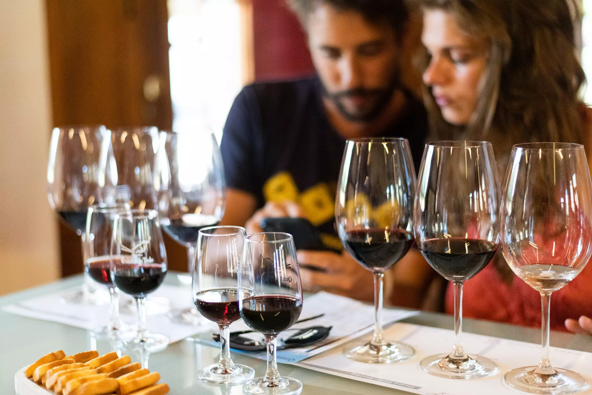 A set of glasses filled with wine are set out for a tasting by a couple, who are visible in the background