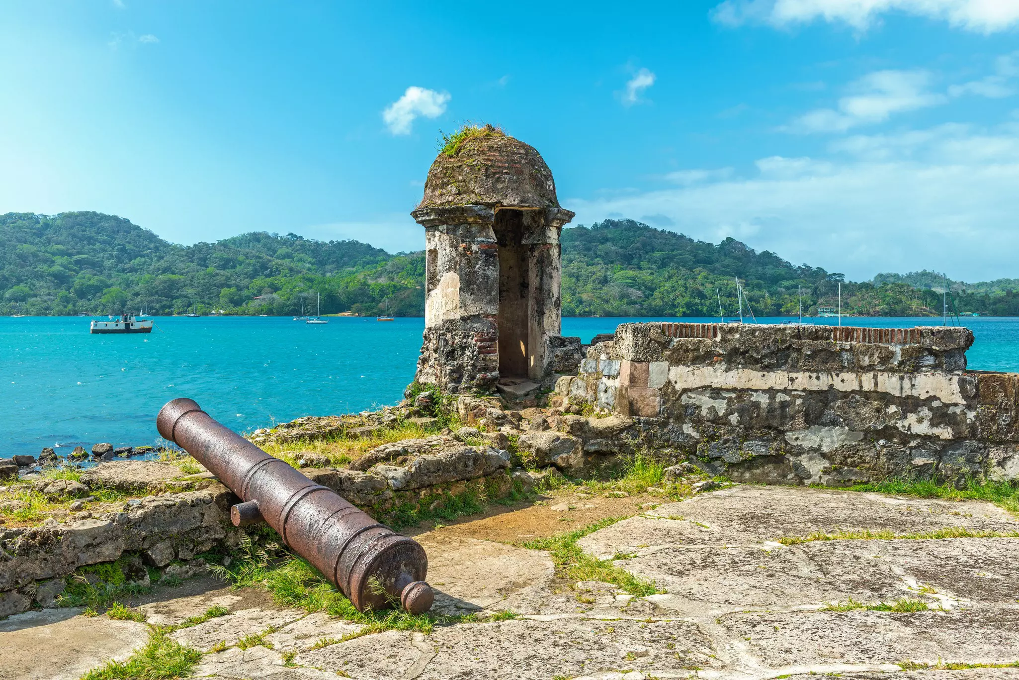 A large iron cannon faces out to see leaning on the ruined walls of an ancient fortress guarding a waterway.