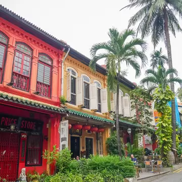 Row of red and yellow two-storey shophouses with wooden shutters and palm trees beside a paved street with cafe tables.