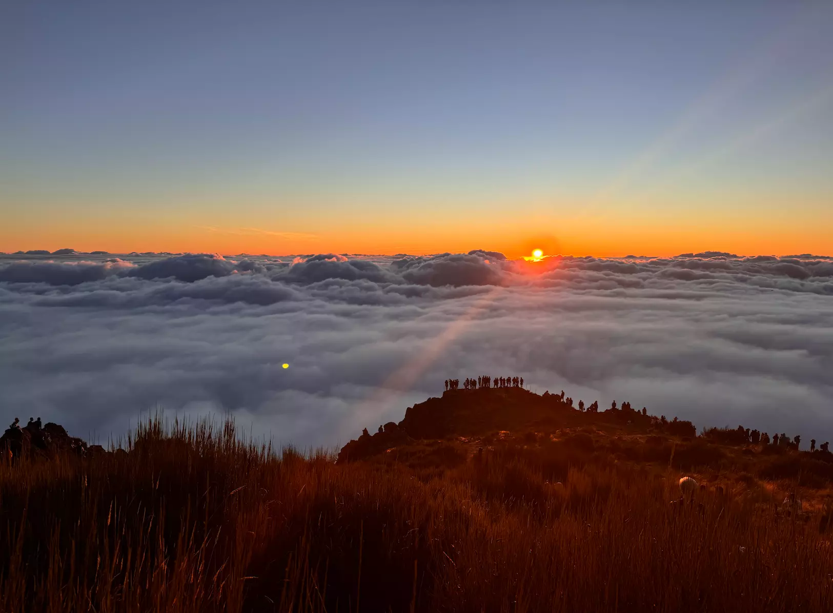 People in silhouette at the edge of a viewpoint watch the sun rise above a layer of cloud
