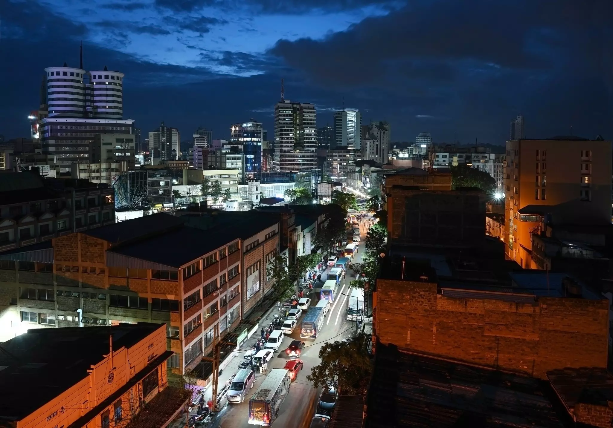 A view looking down from a building of a city street at night, illuminated and filled with cars. The towers of the city skyline are seen in the distance.