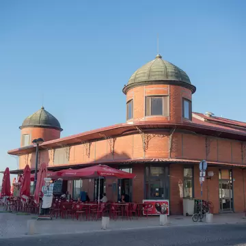 A long red-brick building with two green-domed corner towers, red umbrellas and cafe seating outside under a clear sky.
