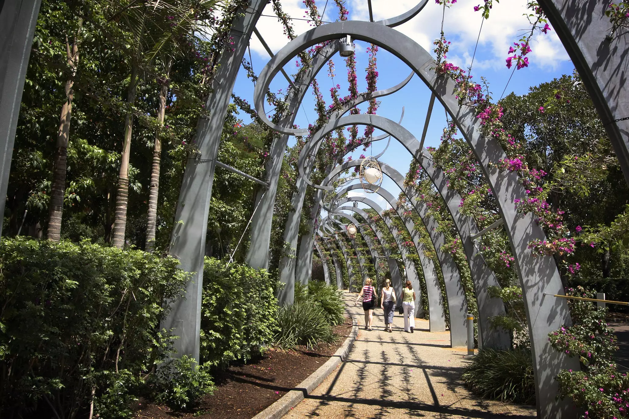 Three women walk along a stone path covered in modern, arched trellis with flowers above on a sunny day.