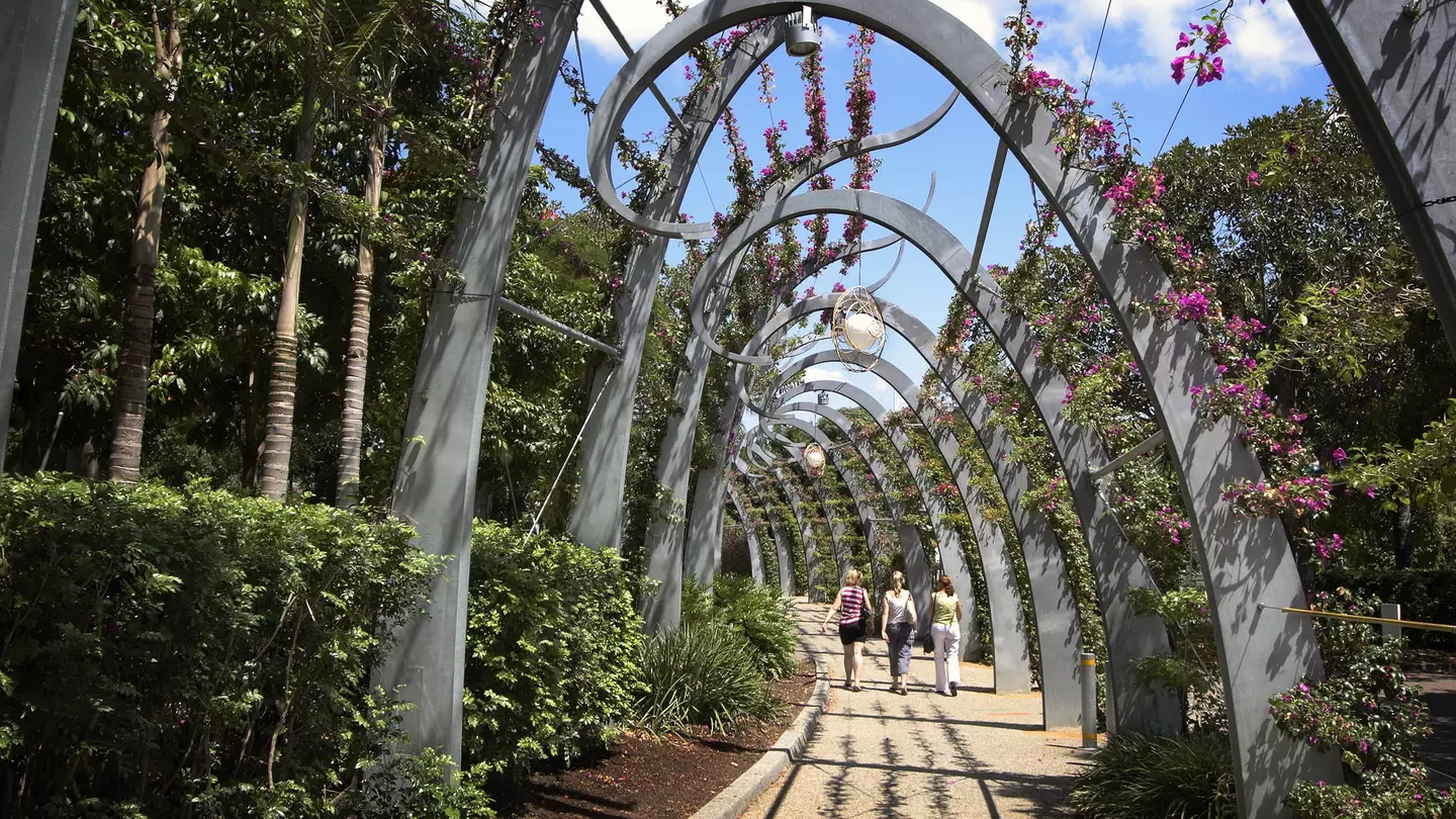 Three women walk along a stone path covered in modern, arched trellis with flowers above on a sunny day.