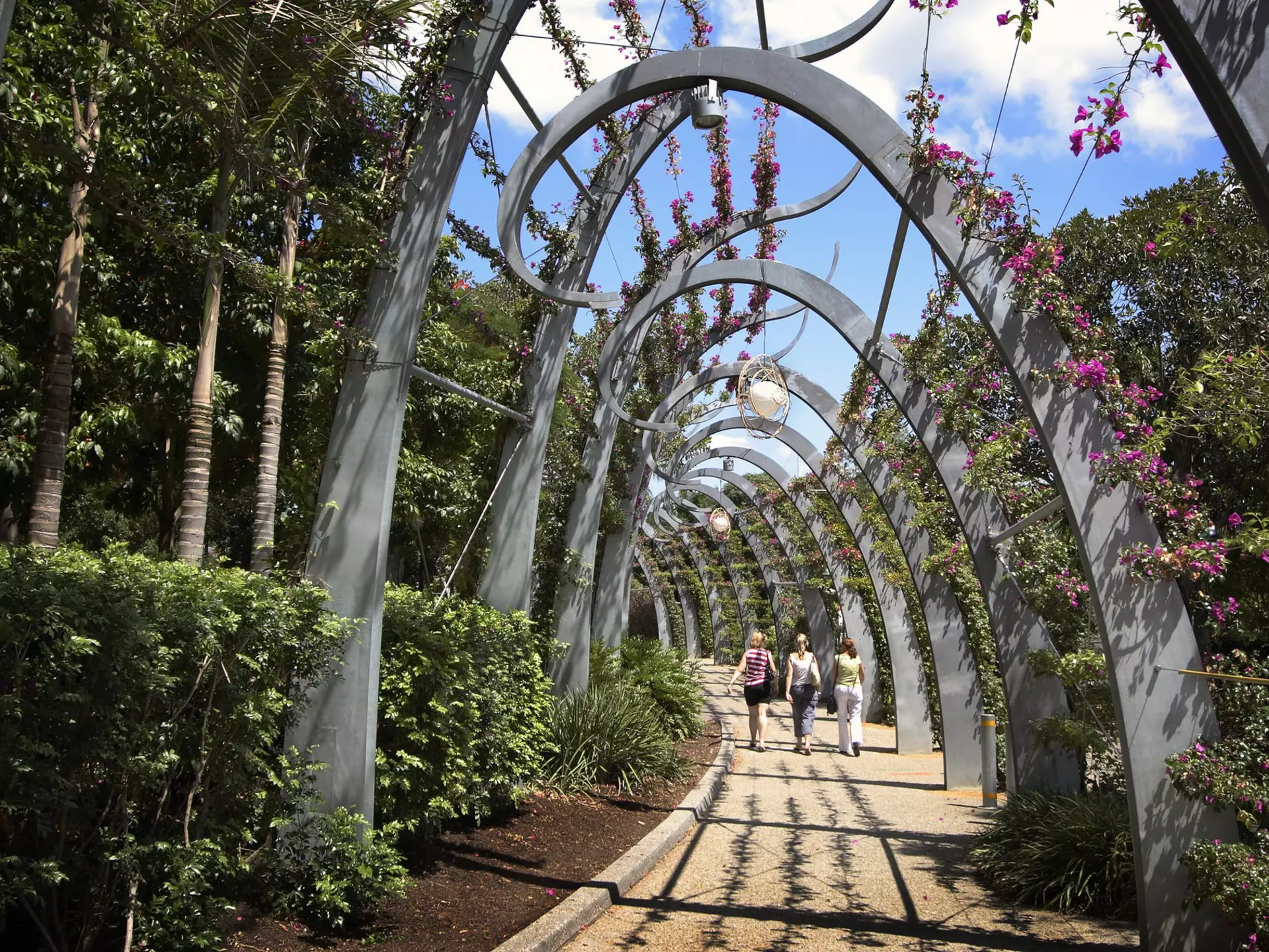 Three women walk along a stone path covered in modern, arched trellis with flowers above on a sunny day.