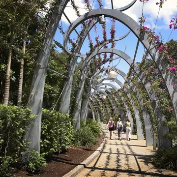 Three women walk along a stone path covered in modern, arched trellis with flowers above on a sunny day.