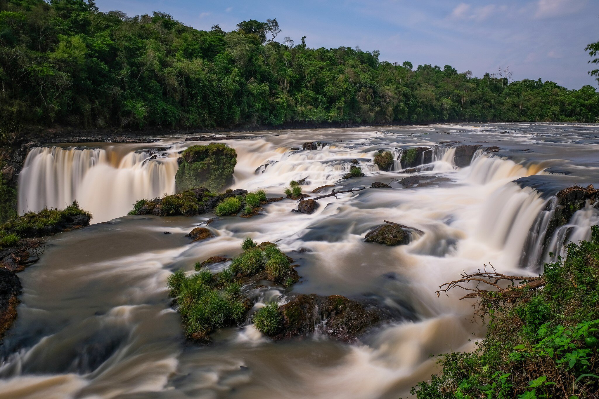 A wide waterfall breaking over a broad rock lip in several curtains, silky from a long exposure, dense forest along the far bank.