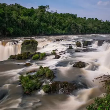 A wide waterfall breaking over a broad rock lip in several curtains, silky from a long exposure, dense forest along the far bank.