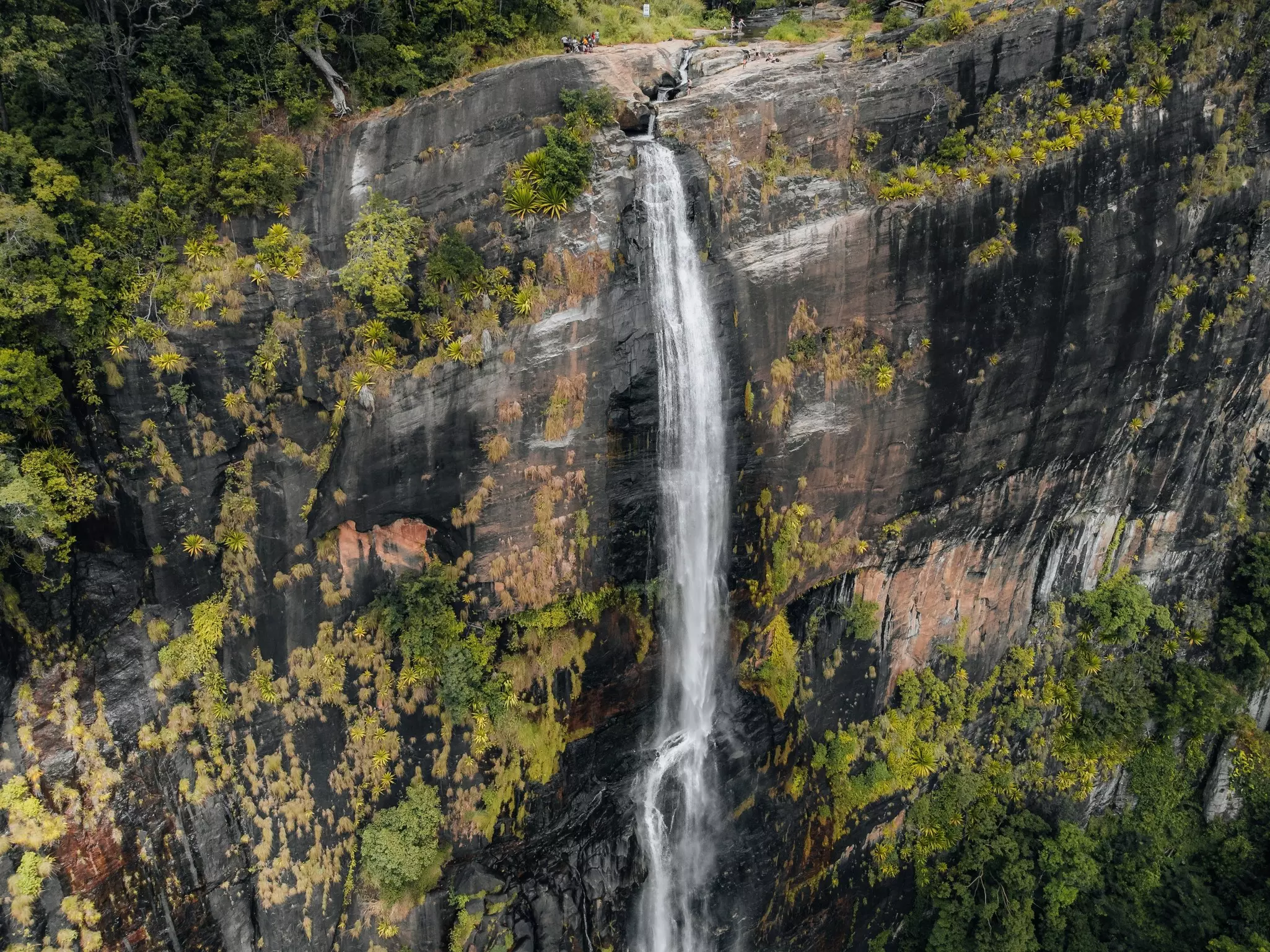 An aerial view of a very tall and narrow waterfall plunging over a cliff. Hikers can be seen at the top of the waterfall.