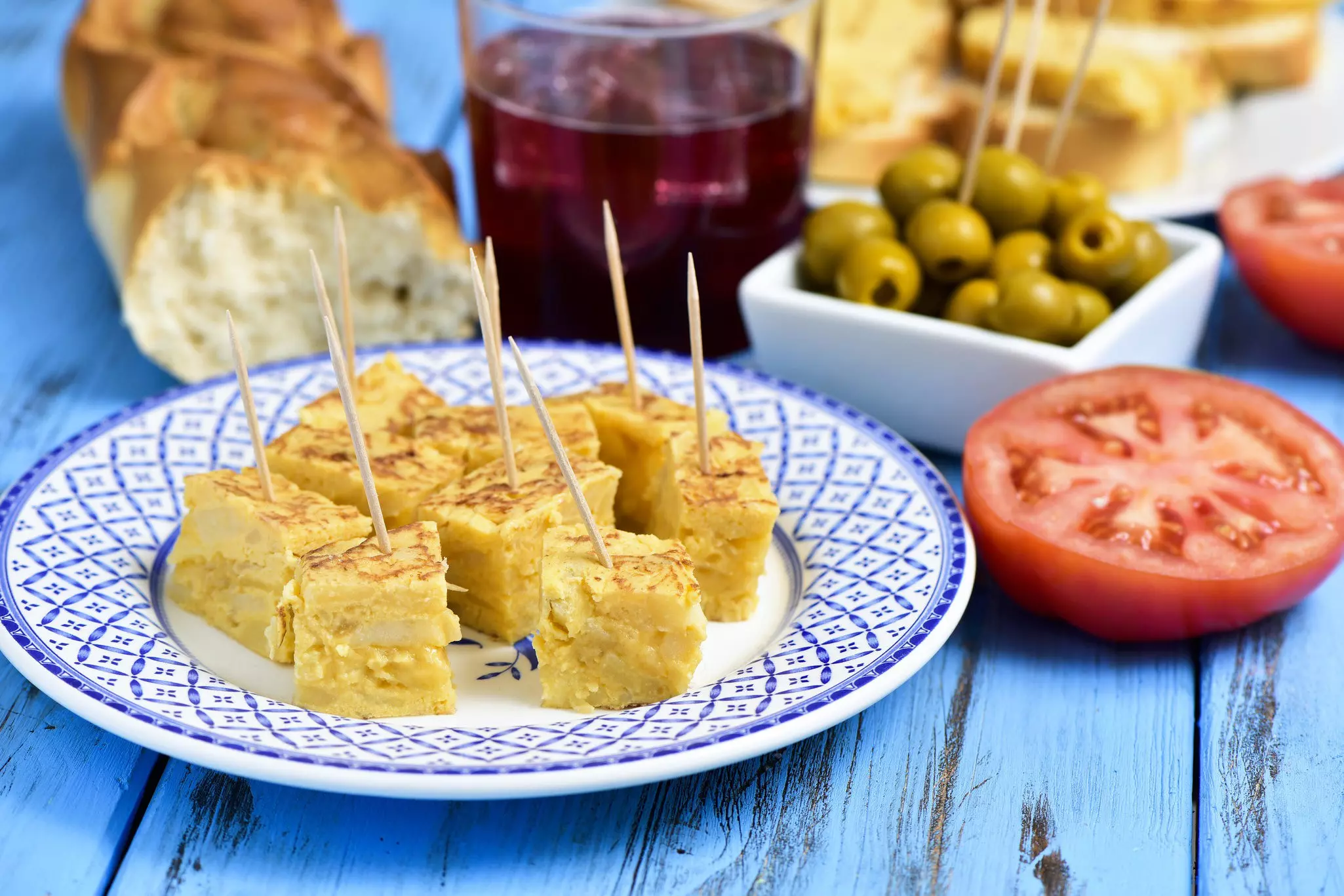 Closeup of a plate with tortilla de patatas, spanish omelet, served as tapas, a glass with tinto de verano, bread and a bowl with olives on a colorful blue wooden table.
