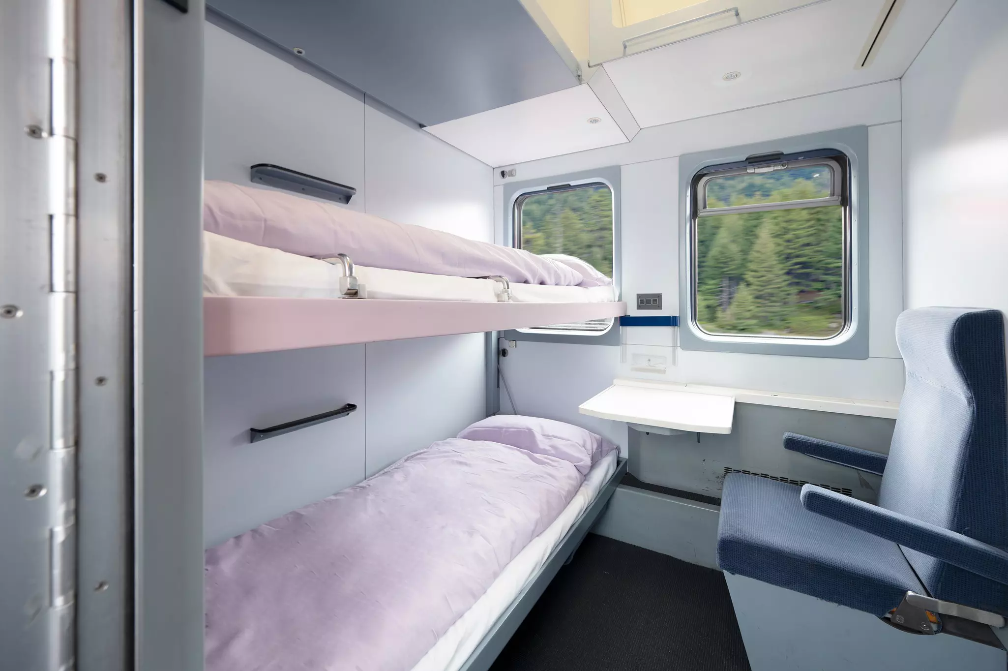 Sleeper car with two narrow bunkbeds and a seat with two windows looking out at trees whizzing past.