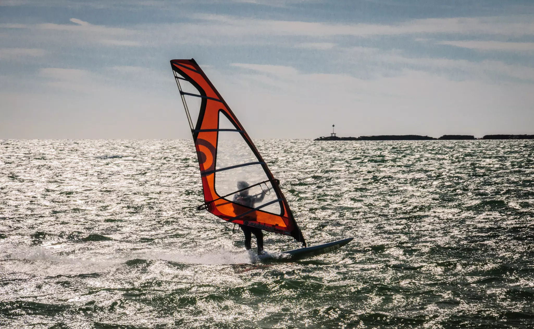 A backlit wind surfer seems skimming across a silver ocean with small waves, with a breakwater in the distance on a bright day.