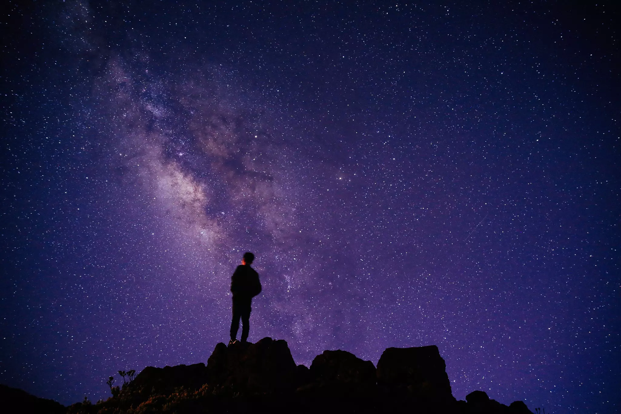 man stands in silhouette looking at the milky way and other stars on a purple night sky