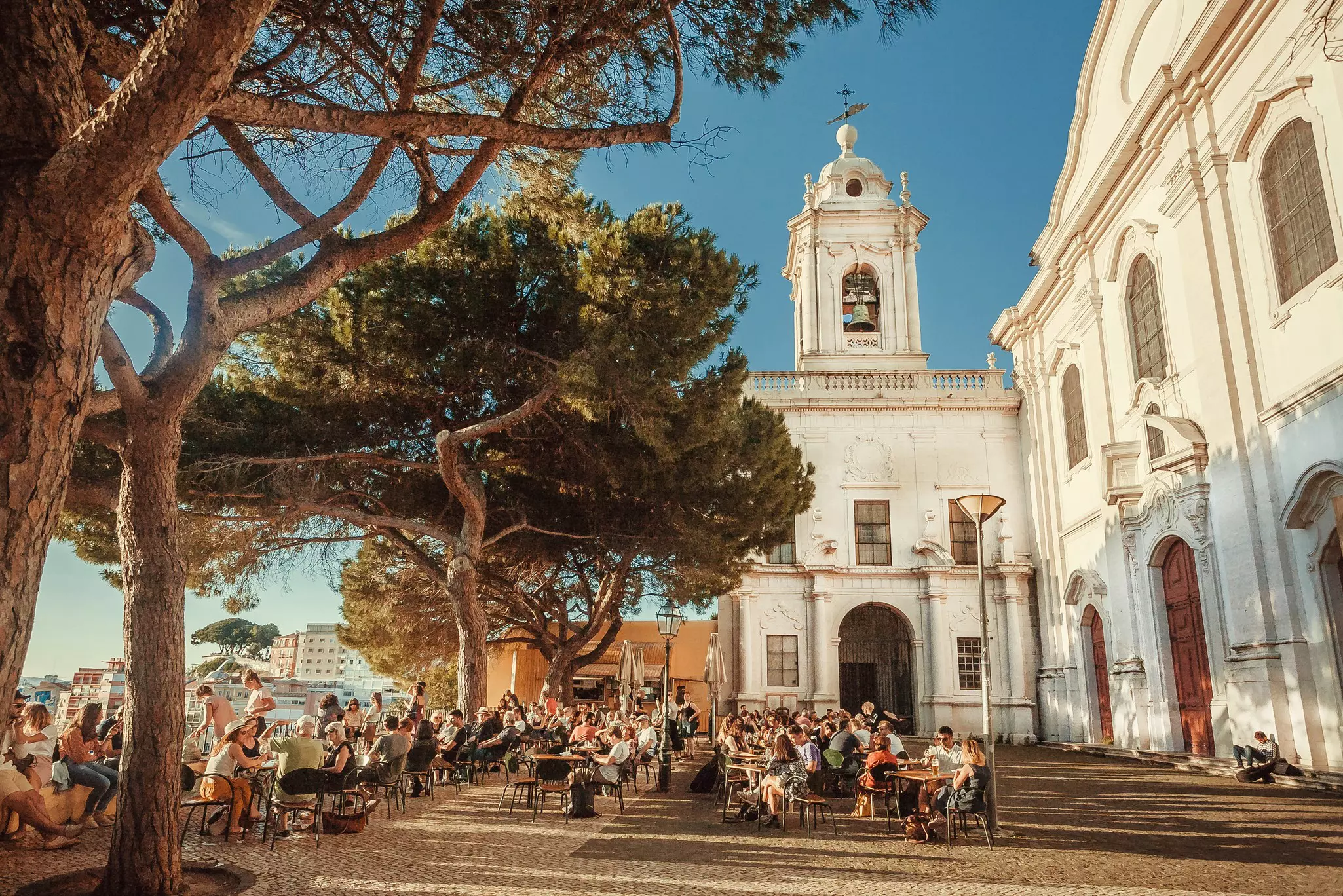 People sit at outdoor tables in a plaza next to a church in a city. Large trees shade the tables.