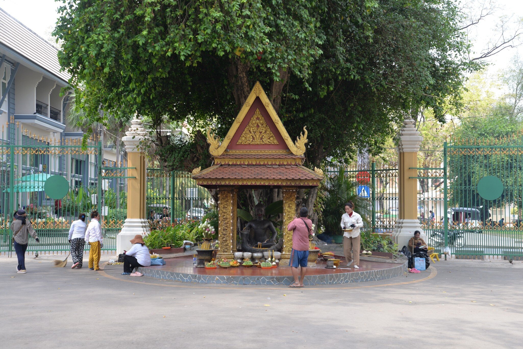 Small gilded pavilion with a dark seated statue under a large tree, people making offerings in front of ornate green gates
