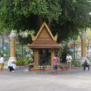 Small gilded pavilion with a dark seated statue under a large tree, people making offerings in front of ornate green gates