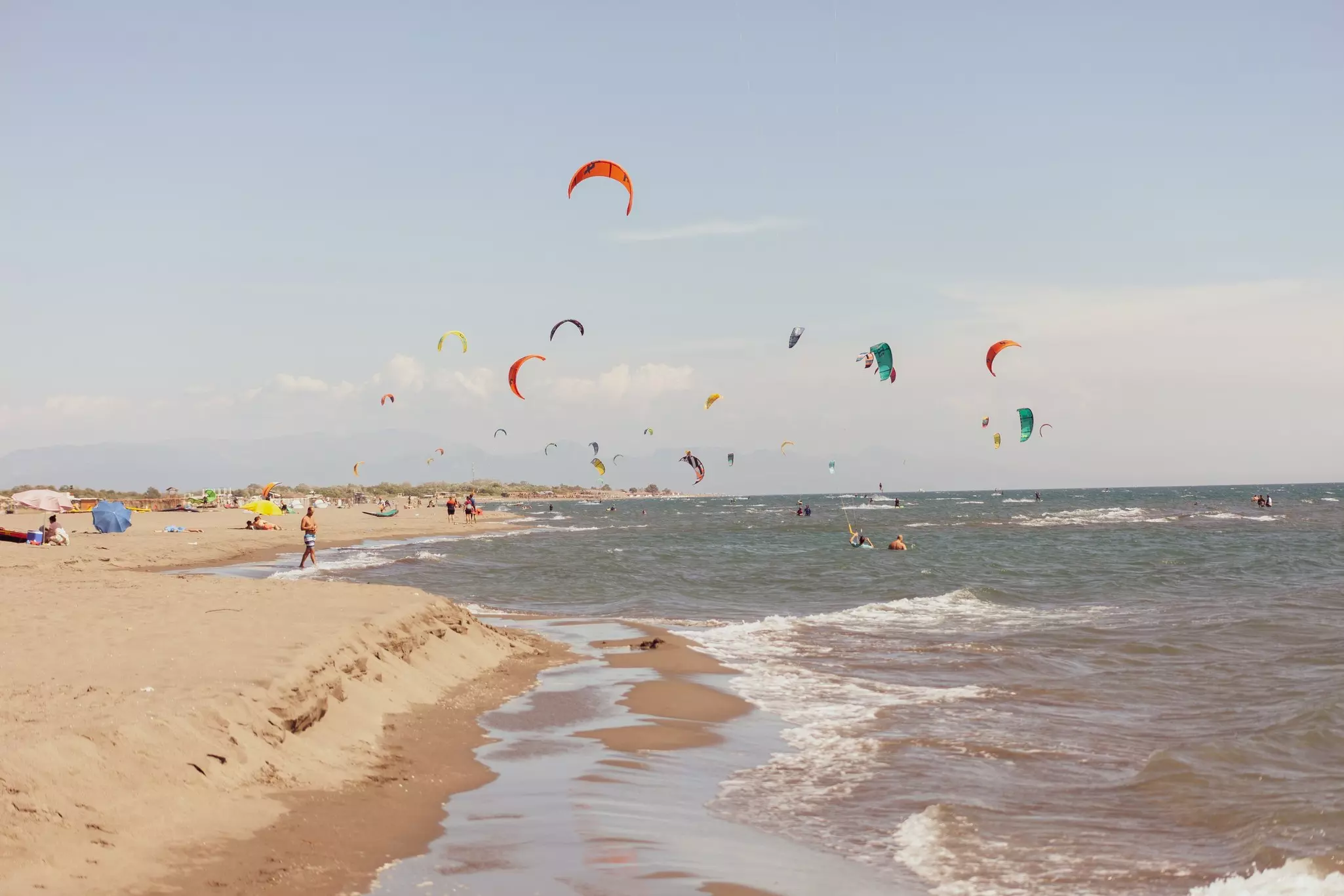 The sails of kitesurfers are seen above a wide beach lapped by white-capped waves.