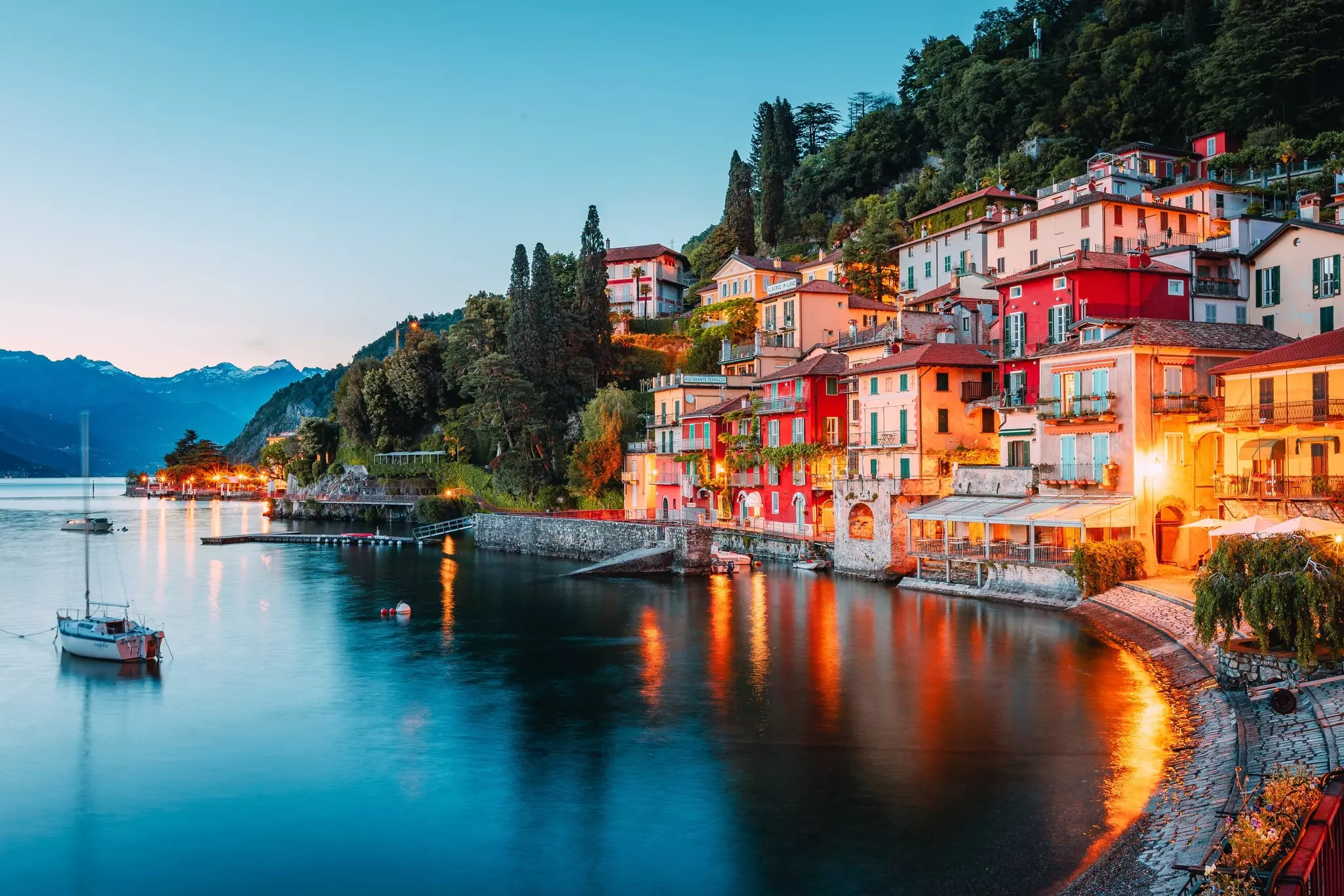 Colorful buildings in a town by a large blue lake are lit up at night, with reflections cast on the water.