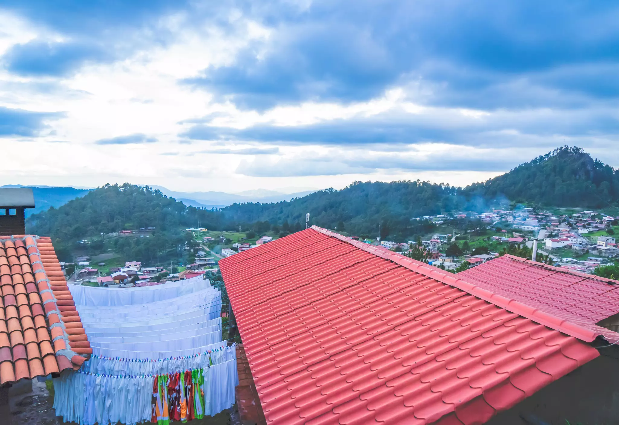 Red roofs of buildings in a mountain village. Green hills lie in the distance, while laundry hangs drying between the buildings.