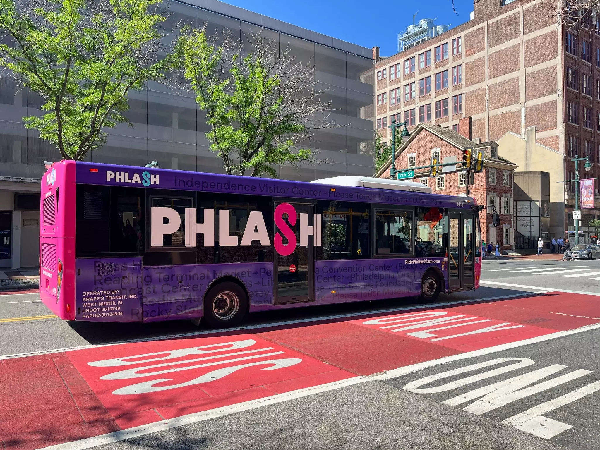 A purple bus with the word "Plash" on it driving down a city street near a red-painted bus-only lane on a sunny day.