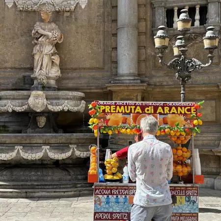A vendor is seen from behind at his colorful stall, which sells juices. The stall is in front of an elaborate fountain on a city street. 